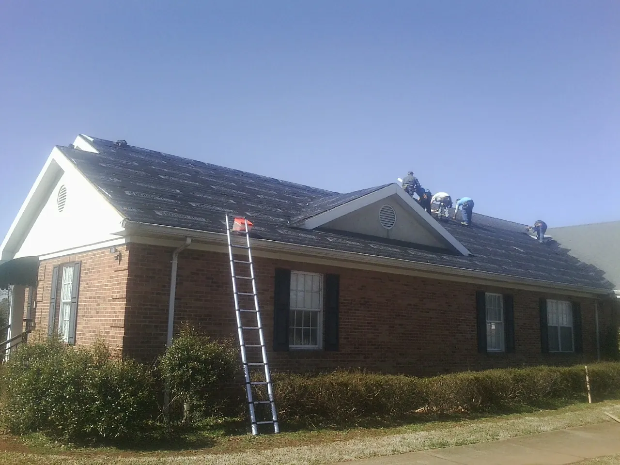 Crew tearing shingles off a brick ranch roof with a ladder set — Emergency Roof Repair work by our crew in De Witt