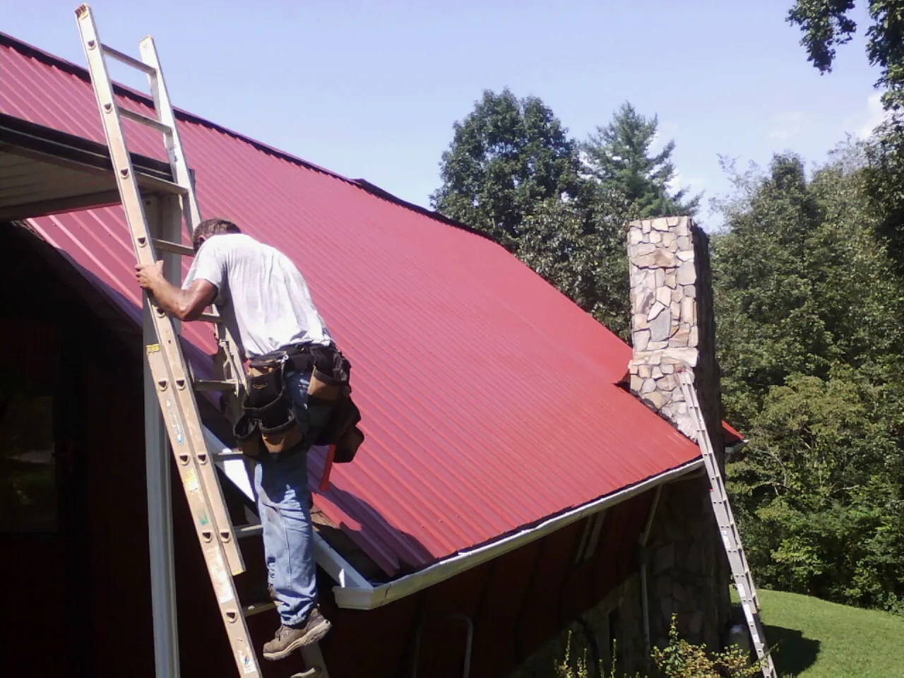 Roofer on a ladder beside a red metal roof and stone chimney — Soffit & Fascia Repair by Roofing Brothers in De Witt